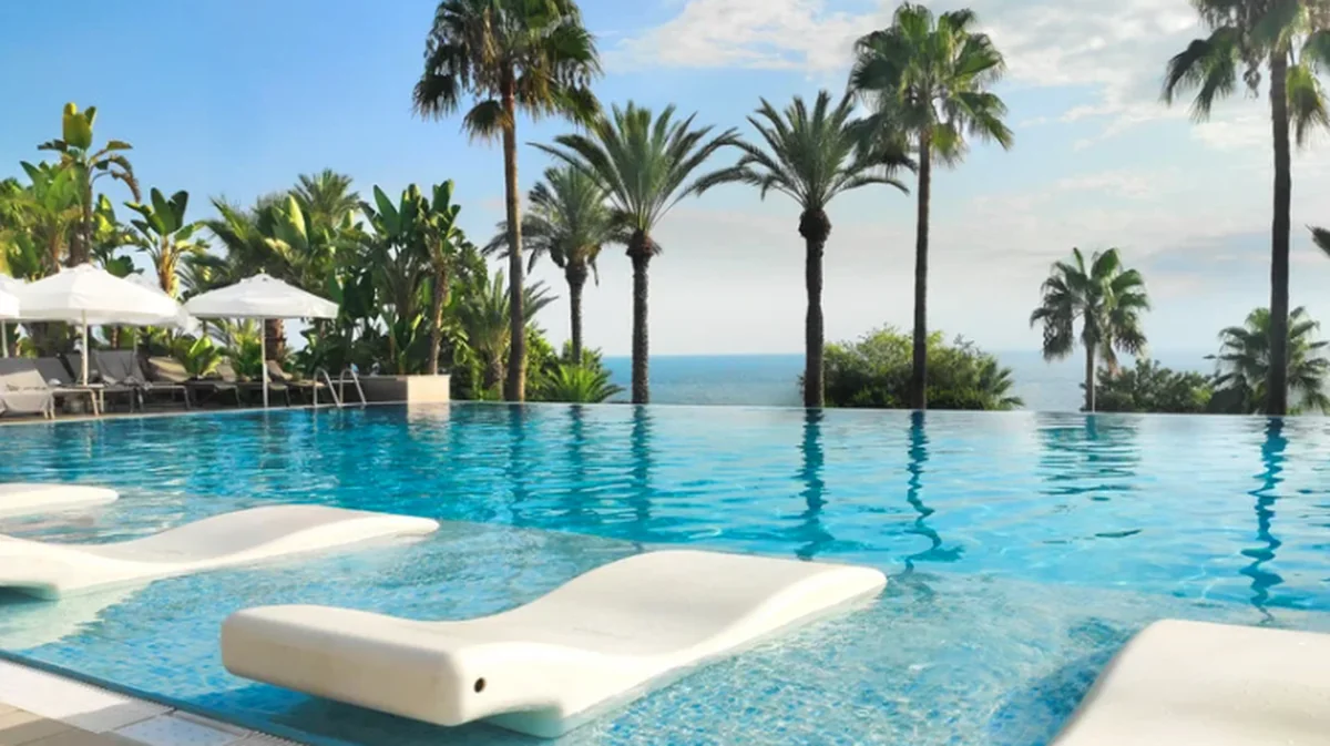 An infinity pool at a luxury resort, lined with floating white loungers and palm trees framing a sea view.